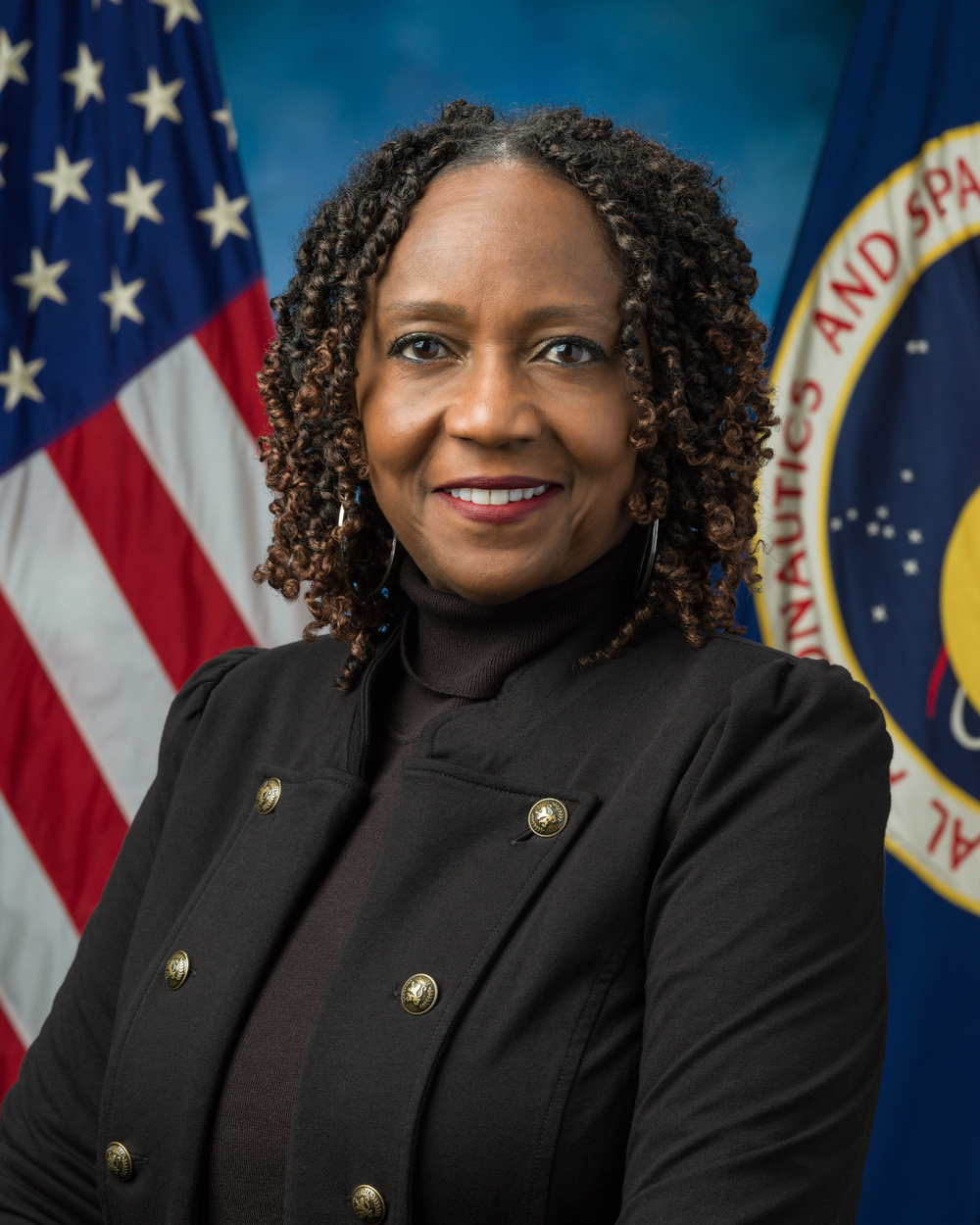 A woman wearing a black blazer smiles in front of a blue background with two flags behind her, a U.S. flag on the left and a NASA flag on the right.
